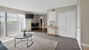 Ein modernes Wohnzimmer mit Flachbildfernseher, rundem Couchtisch, Essbereich mit Stühlen und einer Glasdoppeltür, die auf einen Balkon führt.