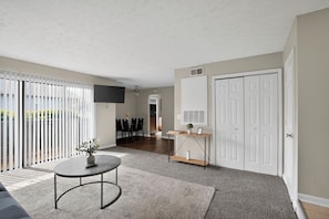 Ein modernes Wohnzimmer mit Flachbildfernseher, rundem Couchtisch, Essbereich mit Stühlen und einer Glasdoppeltür, die auf einen Balkon führt.