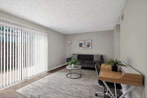 Ein modernes Wohnzimmer mit einer Couch, einem runden Couchtisch mit Pflanze, einem Schreibtisch auf Rollen und einem großen Fenster mit Jalousien.