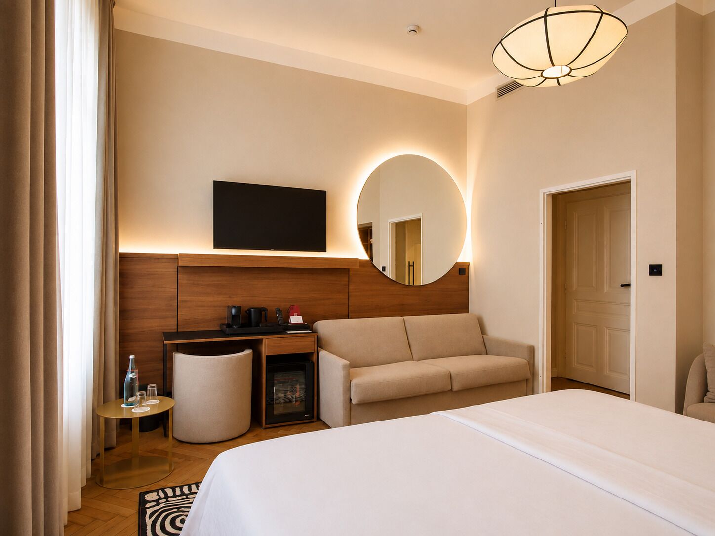 A hotel room with a bed, a sofa, a desk with a flat-screen TV, a mirror, and a round mirror.
