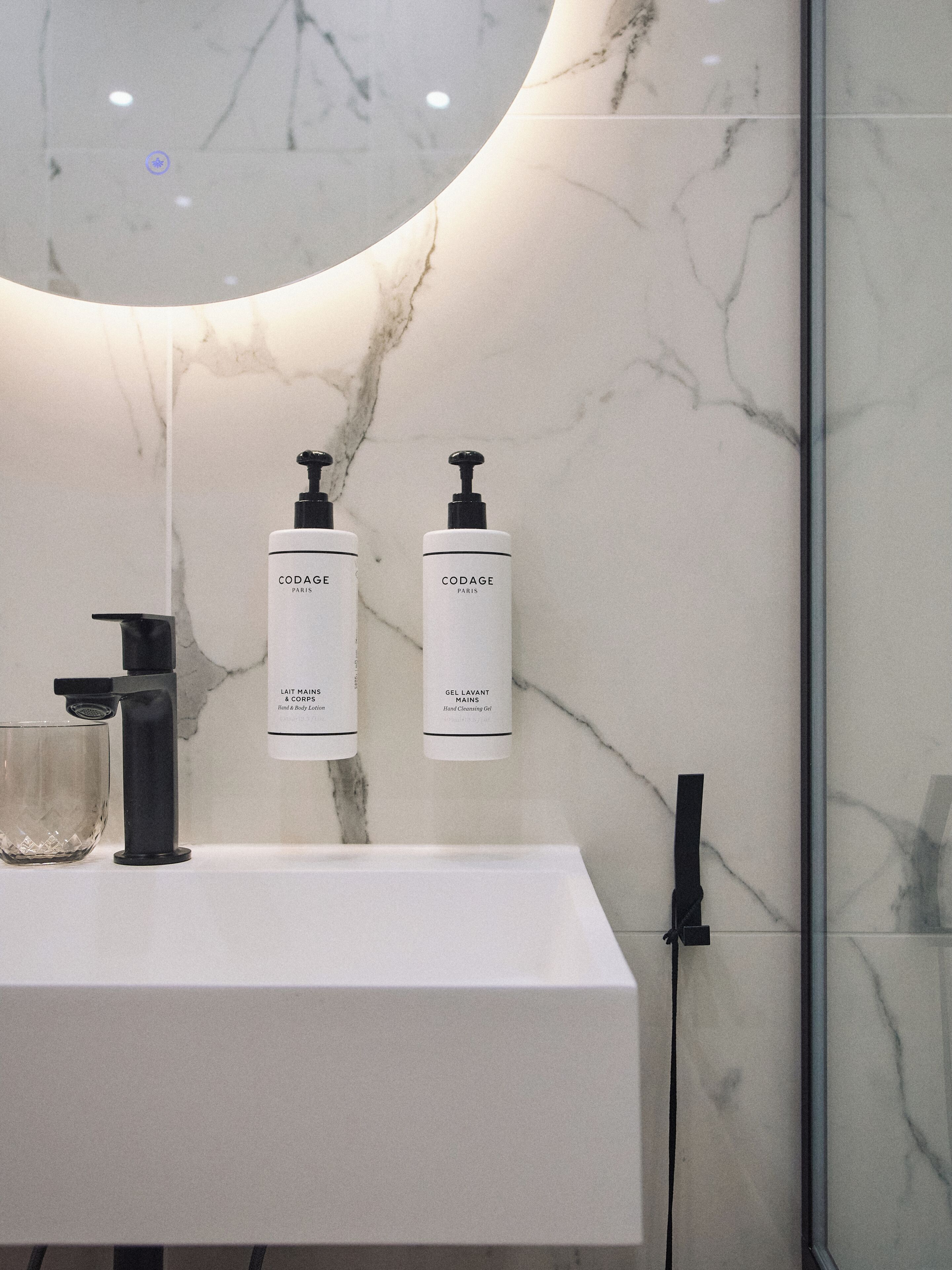 A bathroom with a white marble sink, two bottles of Codage hand cleansing gel, and a black faucet.