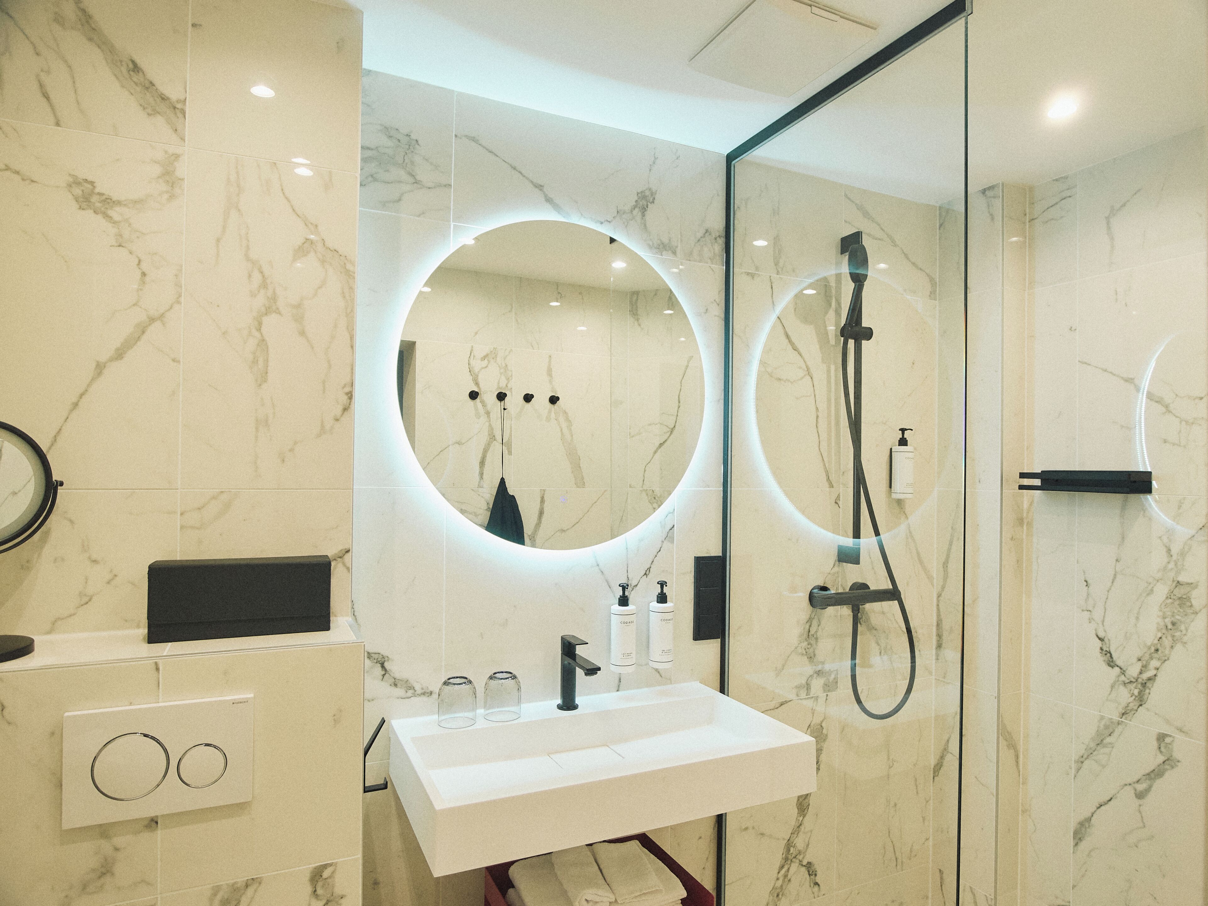 A modern bathroom with a white sink, a glass-enclosed shower, and a circular mirror with built-in lighting.