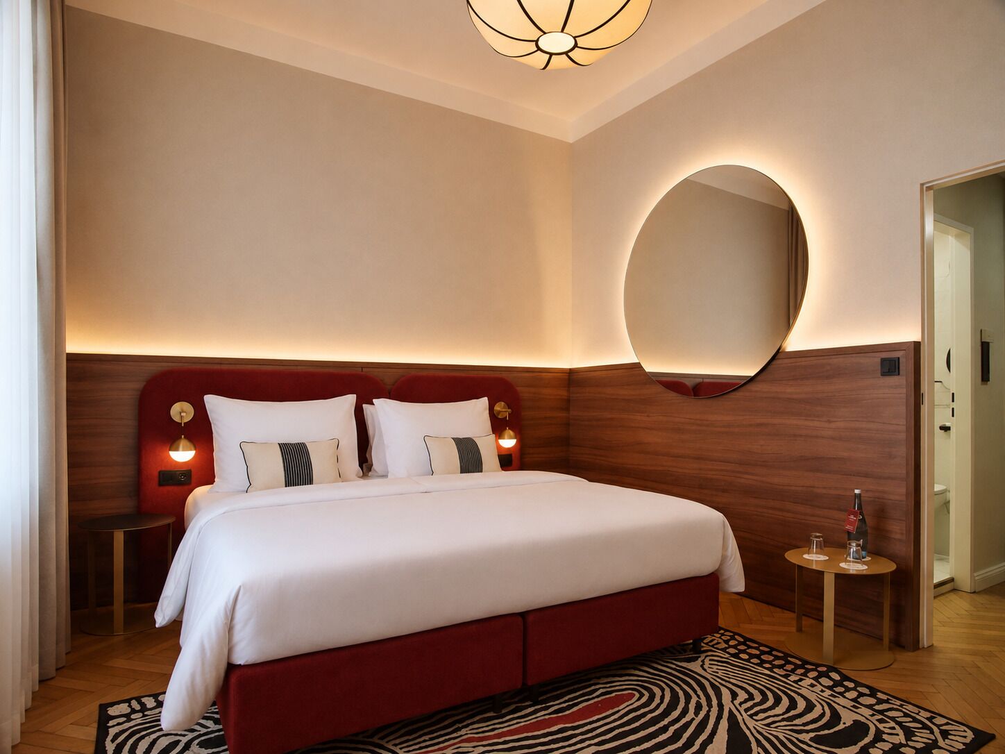 A hotel room with a large bed, a round mirror, a small table with a bottle, and a red headboard.