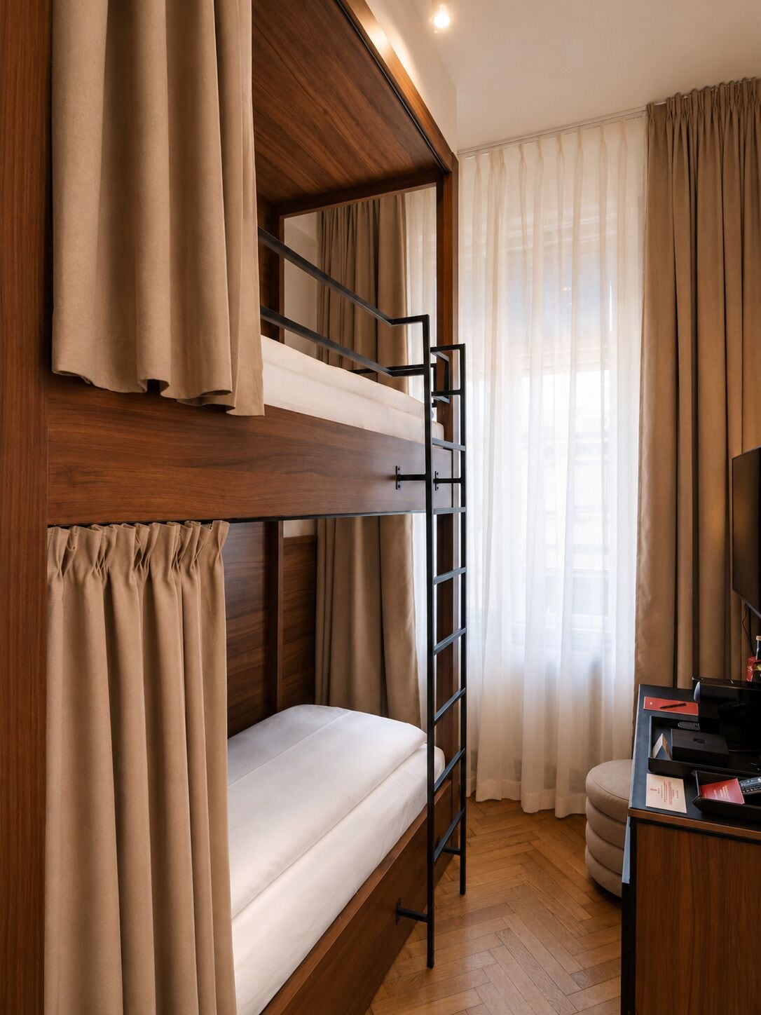 A bunk bed with a ladder, a desk with a computer, and a television in a room with wooden flooring and curtains.