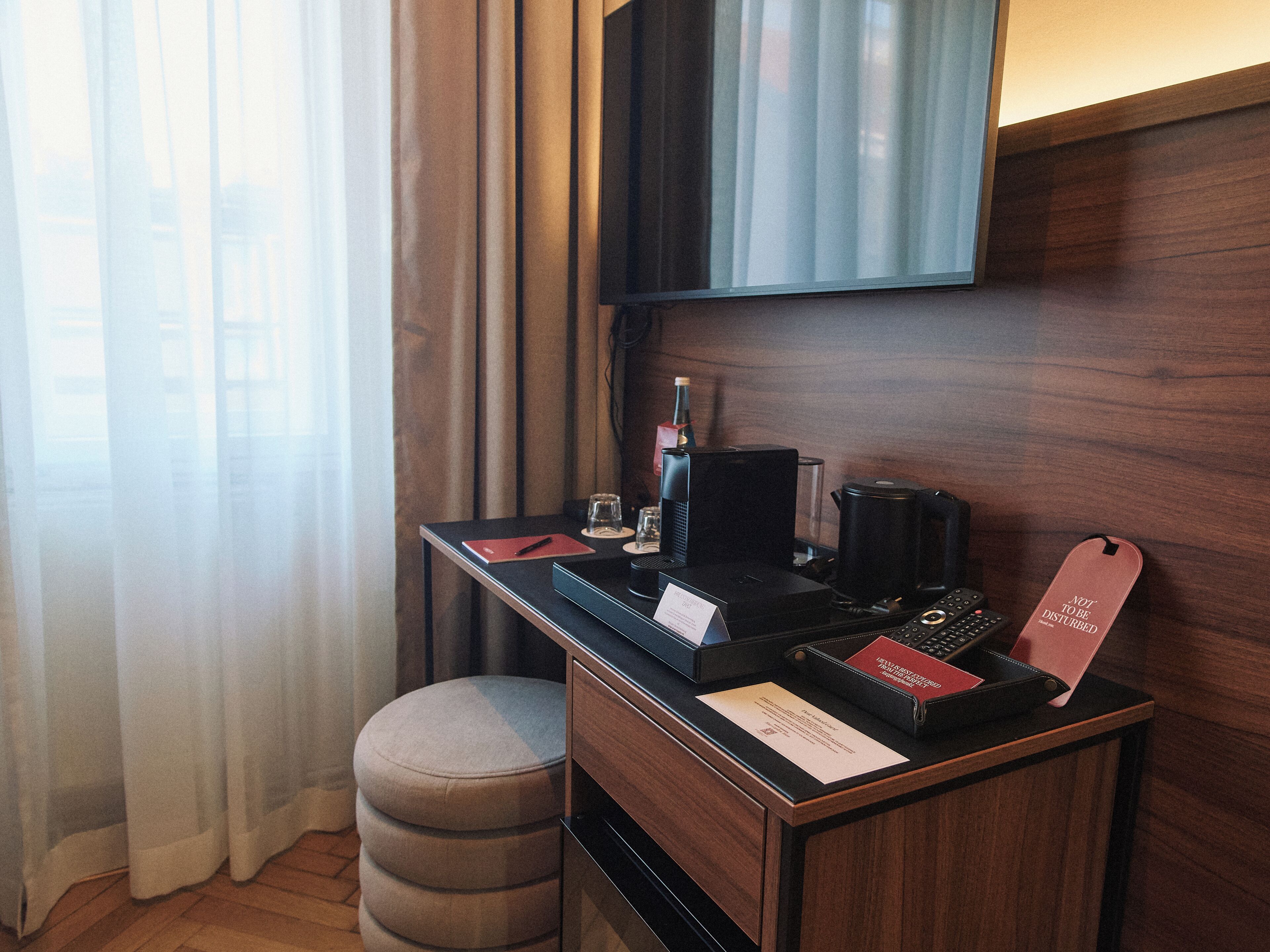 Quarto de hotel com uma escrivaninha, uma cadeira, televisão na parede e uma janela com cortinas.