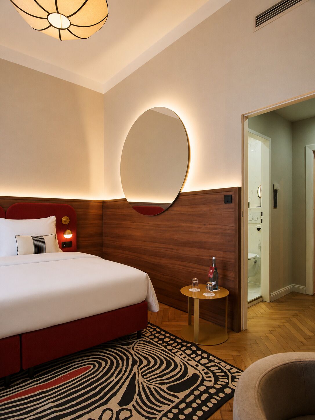 Camera d'albergo con un letto grande, uno specchio rotondo, un tavolino con una bottiglia e un tappeto decorato.