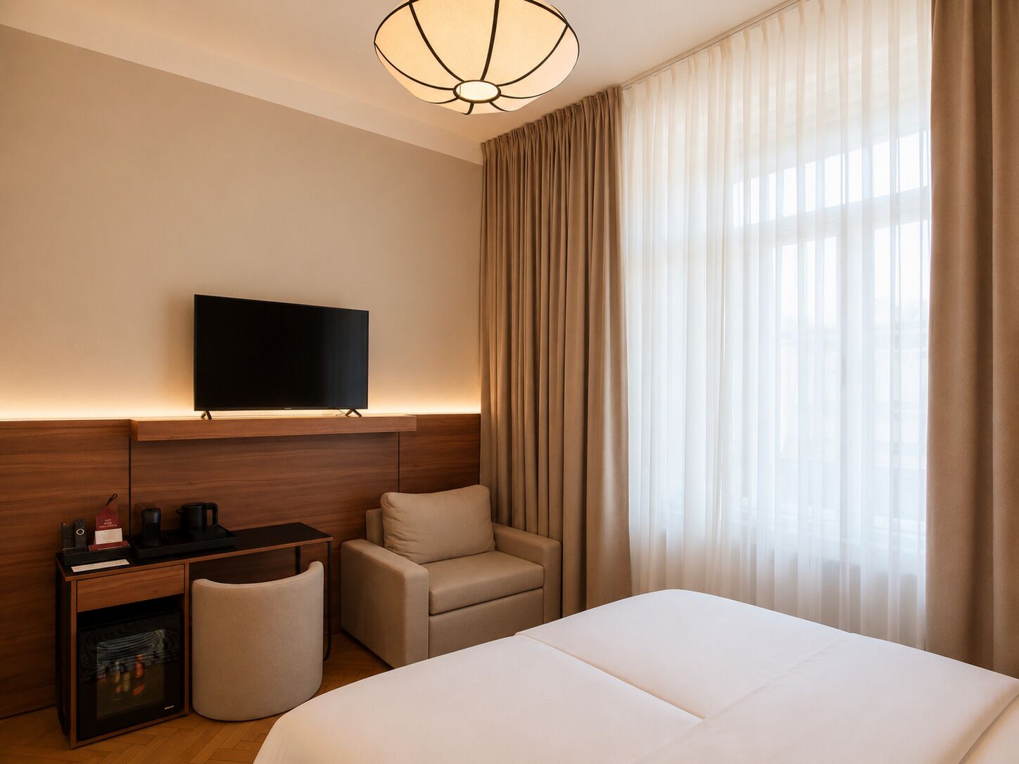 A hotel room with a bed, a desk with a flat-screen TV, a chair, and a large window with curtains.