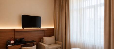 A hotel room with a bed, a desk with a flat-screen TV, a chair, and a large window with curtains.