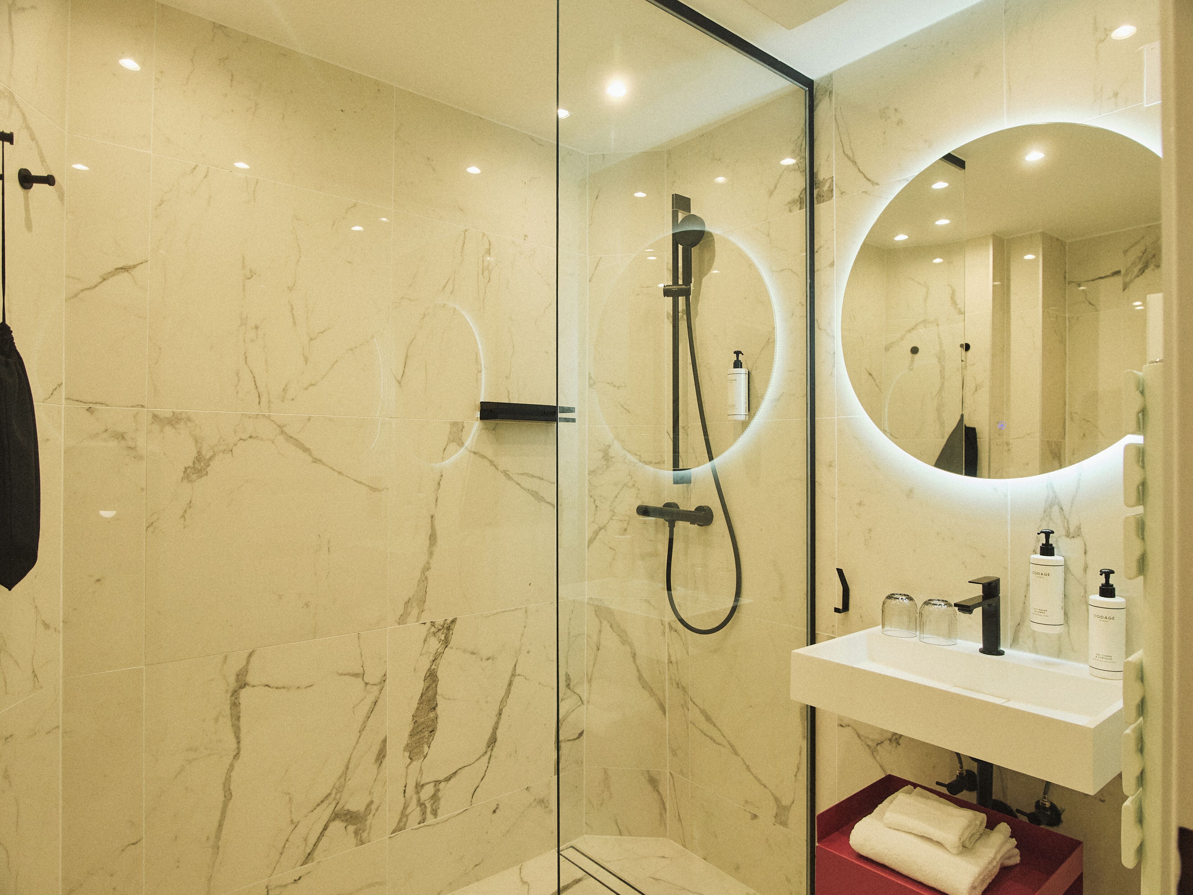 Un baño con ducha de vidrio, lavabo blanco con artículos de aseo y espejo con luz circular.