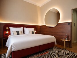 A hotel room with a large bed, a round mirror, a small table with a bottle, and a patterned rug.