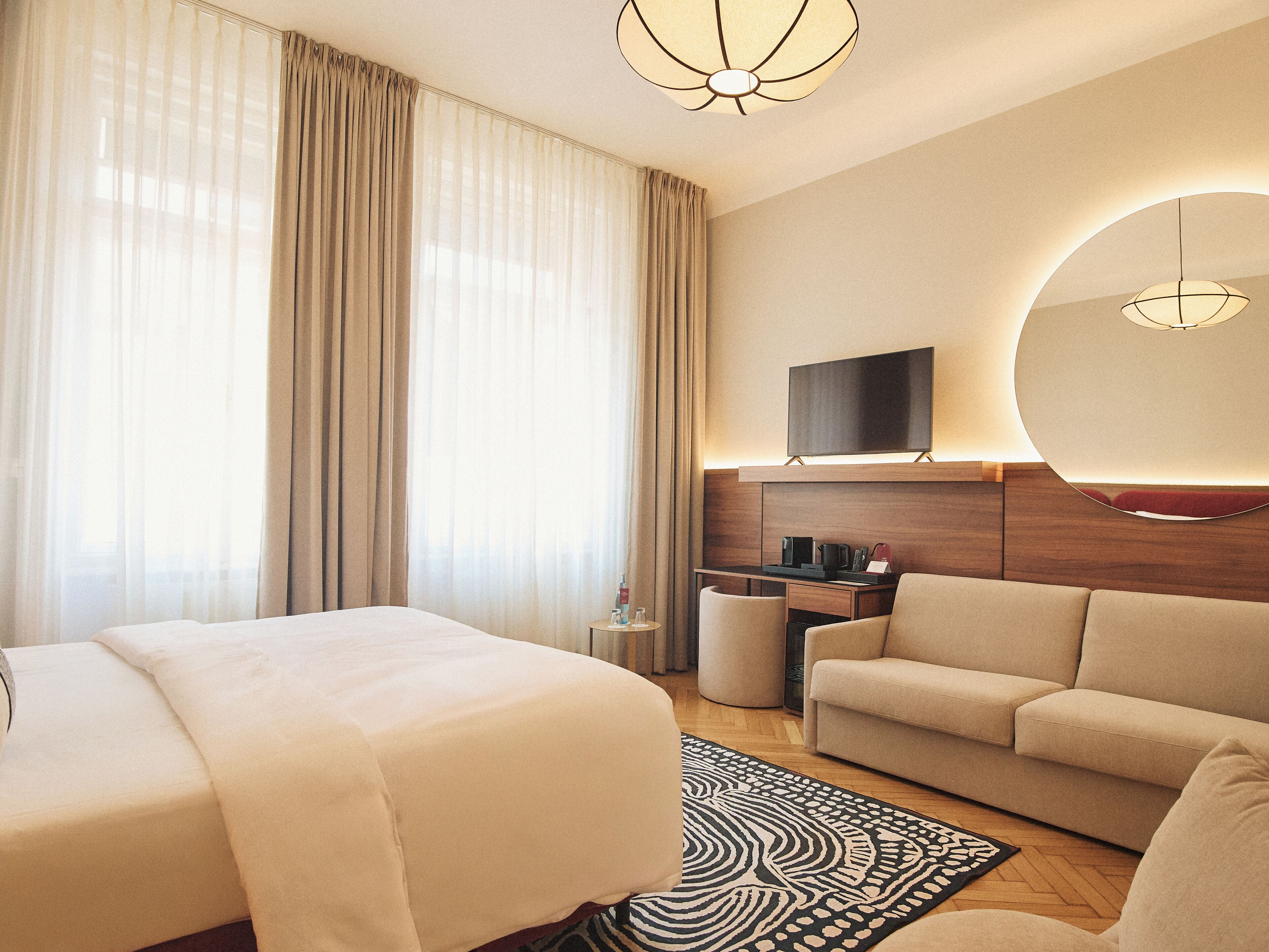 Una habitación de hotel con cama, sofá, televisión y un ventanal con cortinas.