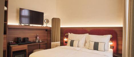 A hotel room with a bed, a TV, a bedside table with a lamp, and a minibar.