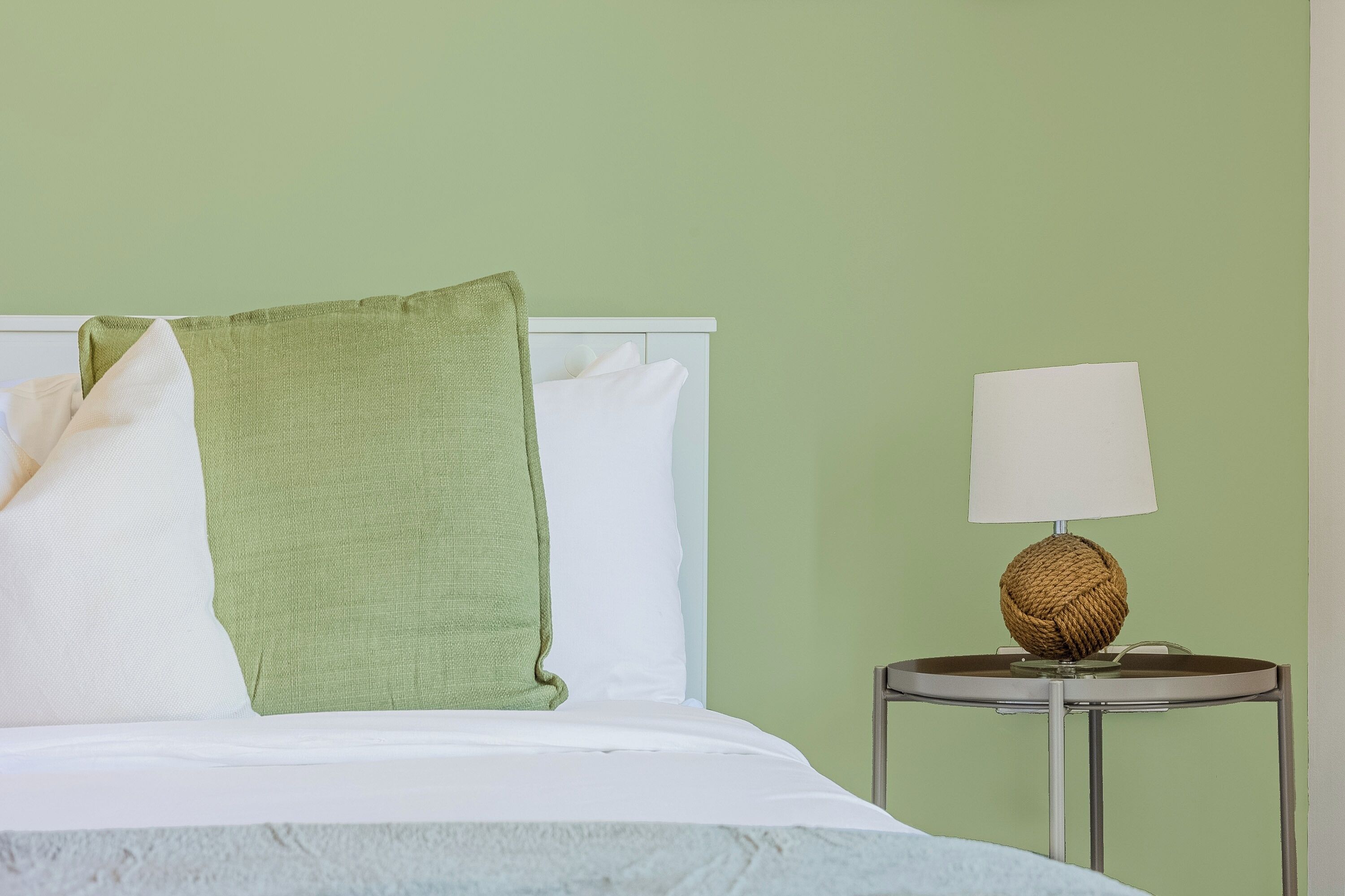 A bed with white linens, a green throw pillow, and a round table with a lamp beside it.
