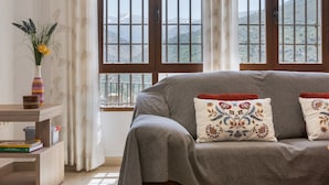 A room with a grey sofa, a wooden side table, a vase with flowers, and a window with a view of mountains. - Apartment 'With Fireplace' with Mountain Views, Wi-Fi and Air Conditioning (Güejar Sierra)