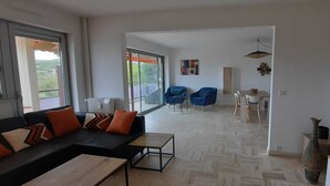Un soggiorno moderno con divano, tavolino da caffè e televisione. C'è un'area pranzo con sedie e un tavolo. La stanza ha ampie finestre e vista sul verde circostante.