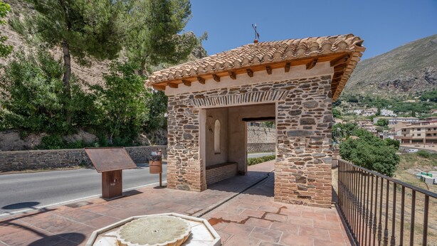 Una estructura de piedra con techo de tejas, un banco y una fuente decorativa de piedra.