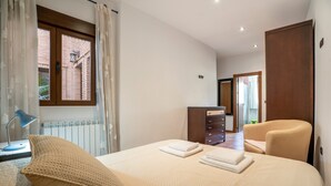 Un dormitorio con cama, una silla, tocador, lámpara y ventana con cortinas.