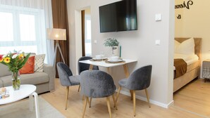 Una sala moderna con sofá, una mesa de comedor con sillas, un televisor de pantalla plana montado en la pared y una cama al fondo.