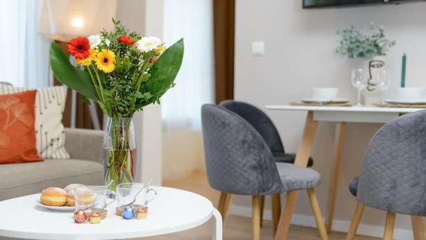 Una sala de estar moderna con una mesa redonda blanca, sillas grises, un jarrón con flores y un televisor montado en la pared.