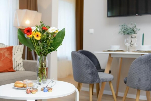 Ein modernes Wohnzimmer mit einem runden weißen Tisch, grauen Stühlen, einer Vase mit Blumen und einem an der Wand befestigten Fernseher.