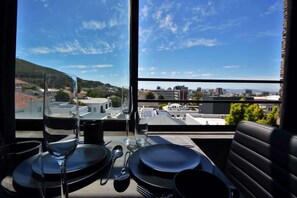 A dining area with a table set for two, a glass of water, and a wine glass. The room has a large window with a view of a cityscape and hills. - Trendy City Pad in Vibrant Location (Cape Town)