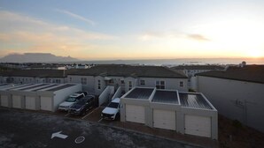 A parking area with several vehicles, including a white van and a black car, in front of a row of buildings with garage doors. - Oceanfront Apartment w/Balcony, Table Mountain (Cape Town)