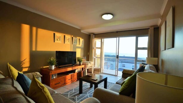 A living room with a TV, sofa, coffee table, and a view of the sea through large windows. - Oceanfront Apartment w/Balcony, Table Mountain (Cape Town)