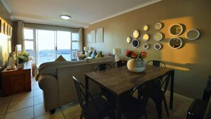 A living room with a sofa, dining table, chairs, and a view through a large window. - Oceanfront Apartment w/Balcony, Table Mountain (Cape Town)