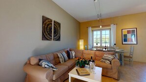 A living room with a sofa, a coffee table with a book and flowers, a dining area with a table and chairs, and a window with curtains. - PASTOUREAU (Sainte-Foy-de-Longas)
