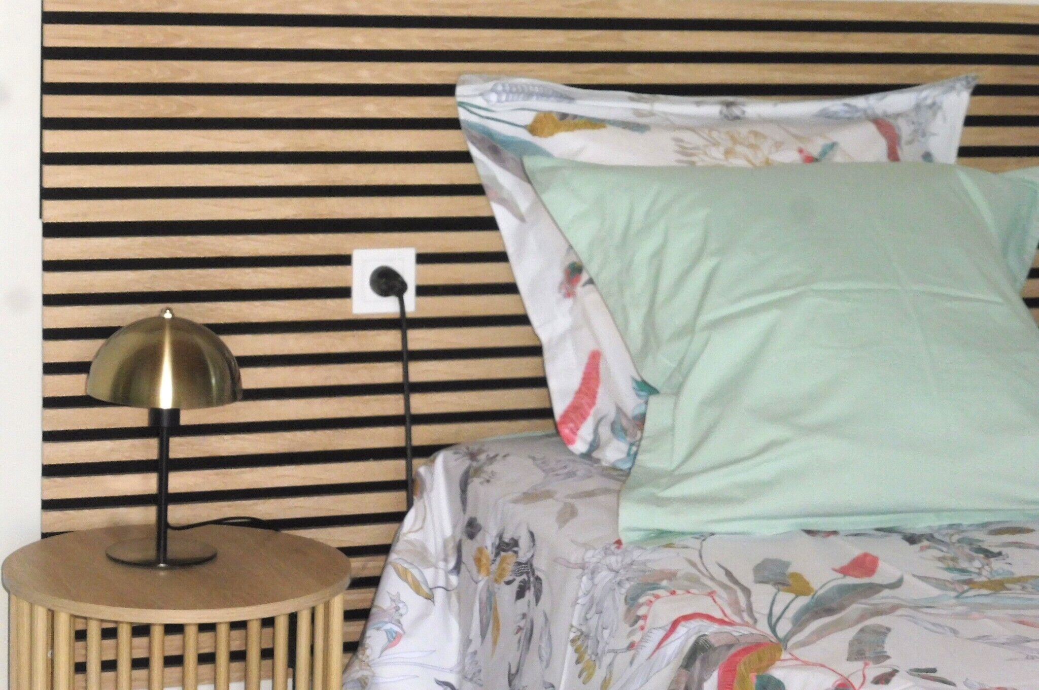 A bed with floral bedding, a round wooden nightstand with a lamp, and a striped wooden headboard.