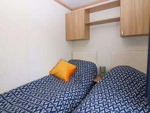 Quarto pequeno e compacto com duas camas, armários de madeira acima e uma luminária em uma mesinha de cabeceira.