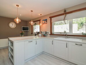 A modern kitchen with white cabinets, pendant lights, a clock, and a window with a view of greenery.