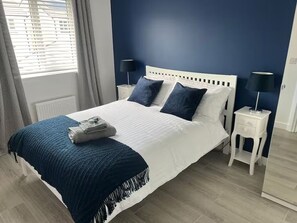 A neatly made bed with a white headboard and dark blue pillows, a blue throw blanket, and a folded towel on the bed. There are two bedside tables with lamps, one on each side of the bed. - St Andrews Golf Getaway - 3.5 miles to Old Course + Private Putting Green (St Andrews)