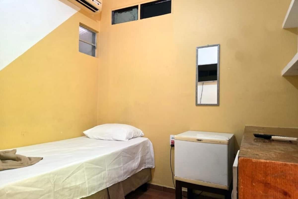 A small, single-bed room with a bed, a small table, a mirror, and a wall-mounted air conditioner.