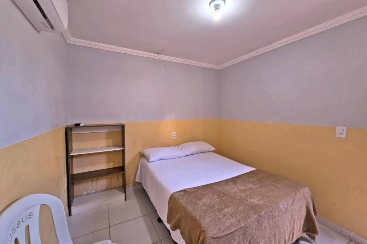 A small, single-bed room with a white bed, a brown blanket, a white chair, a shelf, and an air conditioning unit.