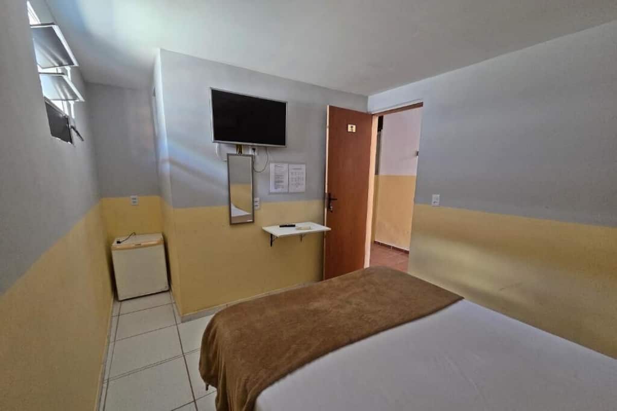 A hotel room with a bed, a television mounted on the wall, a small bedside table, and a door leading to another room.