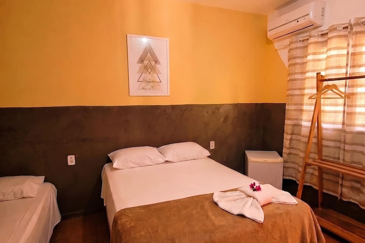 A hotel room with a bed, a bedside table, an air conditioning unit, a towel rack, and a framed picture on the wall.