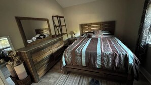A bedroom with a wooden dresser, a large mirror, a bed with patterned bedding, and a nightstand with a lamp.