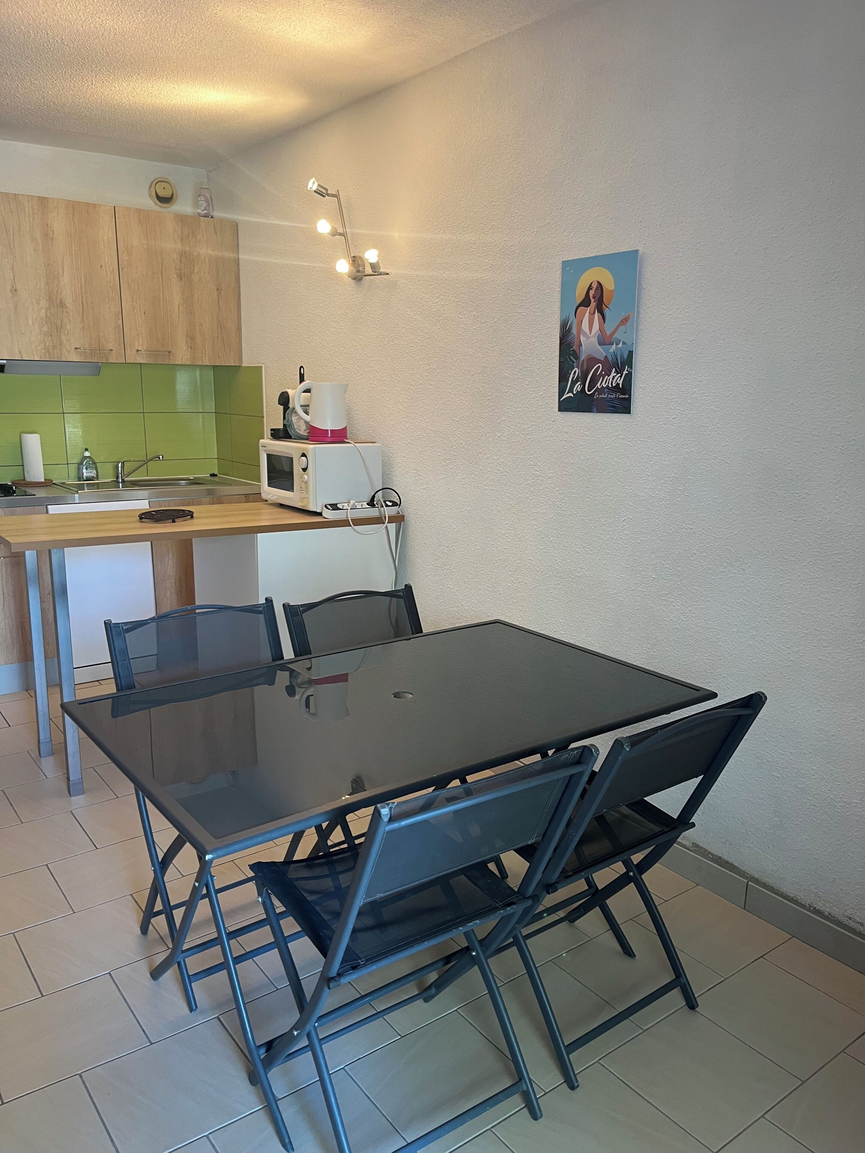 Eine kompakte Küche mit Mikrowelle, Wasserkocher und einer kleinen Spüle. Es gibt einen Esstisch mit Stühlen und ein Poster an der Wand.