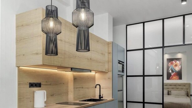 Una cocina moderna con una isla de madera, taburetes negros tipo bar, luces colgantes y un muro divisorio de vidrio.