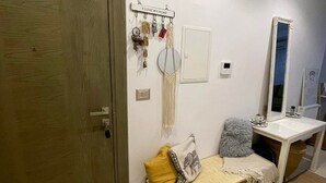 Una stanza con una porta in legno, una panca con cuscini, un portachiavi a parete, uno specchio e un piccolo tavolo bianco con una tazza.