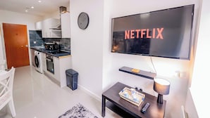 A modern living room with a flat-screen TV displaying 'NETFLIX,' a kitchen with appliances, a dining area, and a clock on the wall. - Town Centre Gem Bright and Spacious 3-Bed (Cardiff)