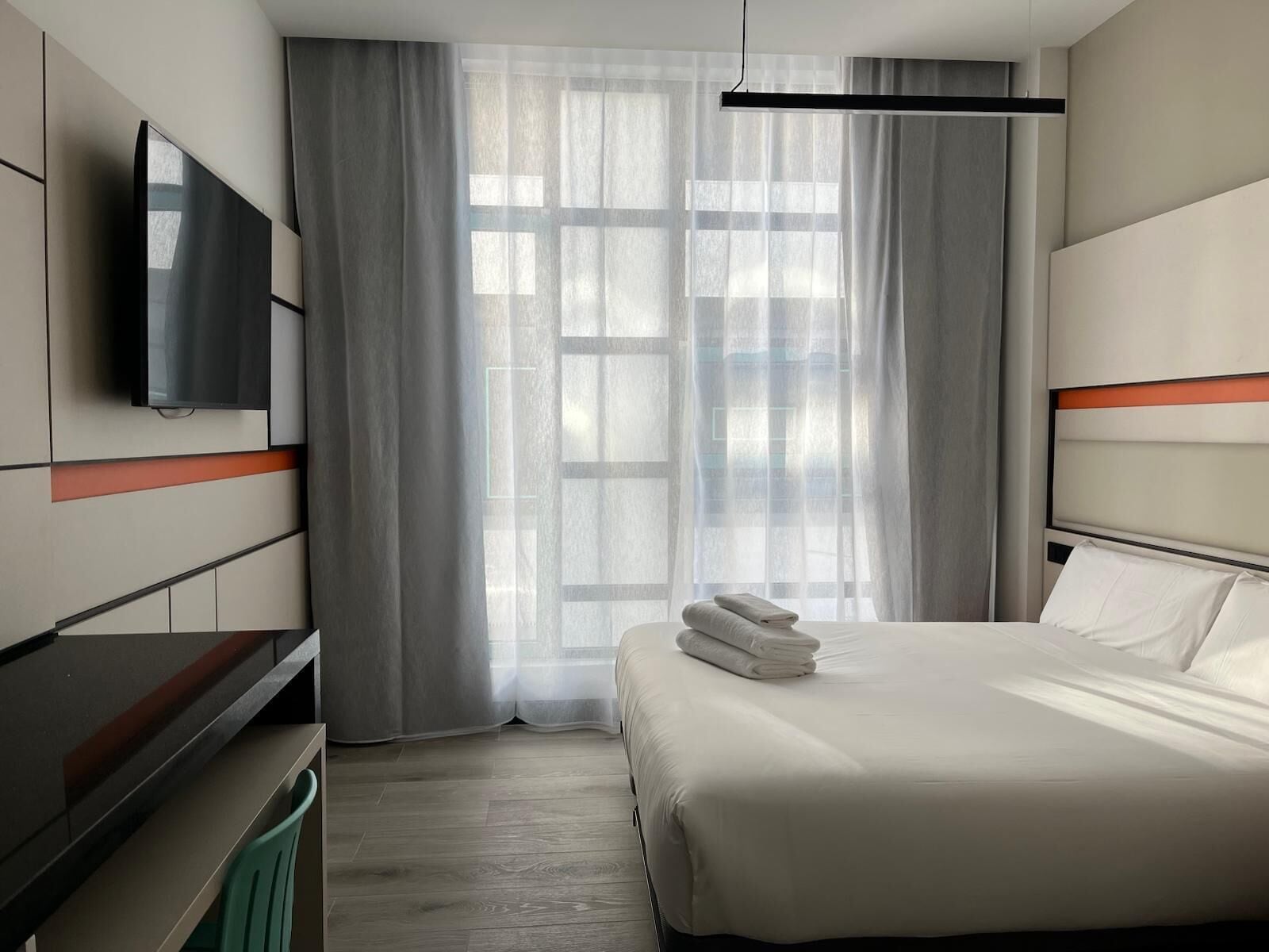 A modern hotel room with a bed, a flat-screen TV, a desk, and a large window with sheer curtains.