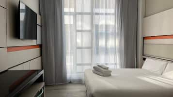 Una habitación de hotel moderna con cama, televisor de pantalla plana, escritorio y ventana grande con cortinas translúcidas.
