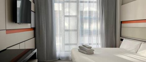 A modern hotel room with a bed, a flat-screen TV, a desk, and a large window with sheer curtains.