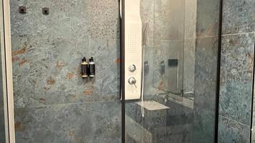 Un baño moderno con ducha envuelta en cristal, paredes de mármol y una zona de lavabo a la vista.