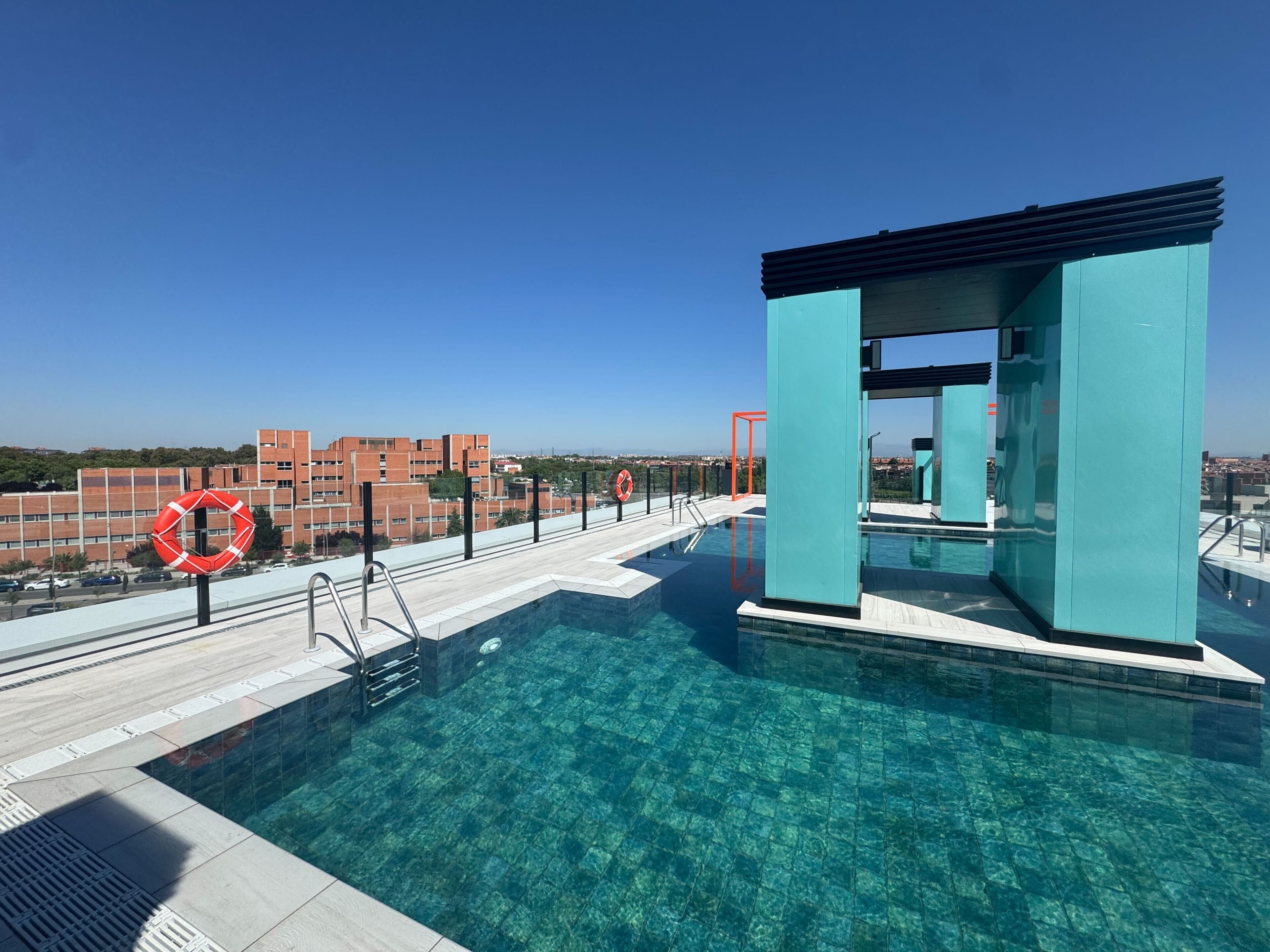 A rooftop swimming pool with clear blue water, a lifebuoy, and a view of buildings in the background.