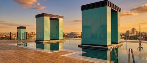 Un área de piscina en la azotea con tres estructuras rectangulares, una terraza de madera y vistas a la ciudad.