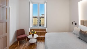 Ein Schlafzimmer mit einem Bett, einem Sessel, einem kleinen Tisch mit Blumen und einem Fenster mit Blick nach draußen.