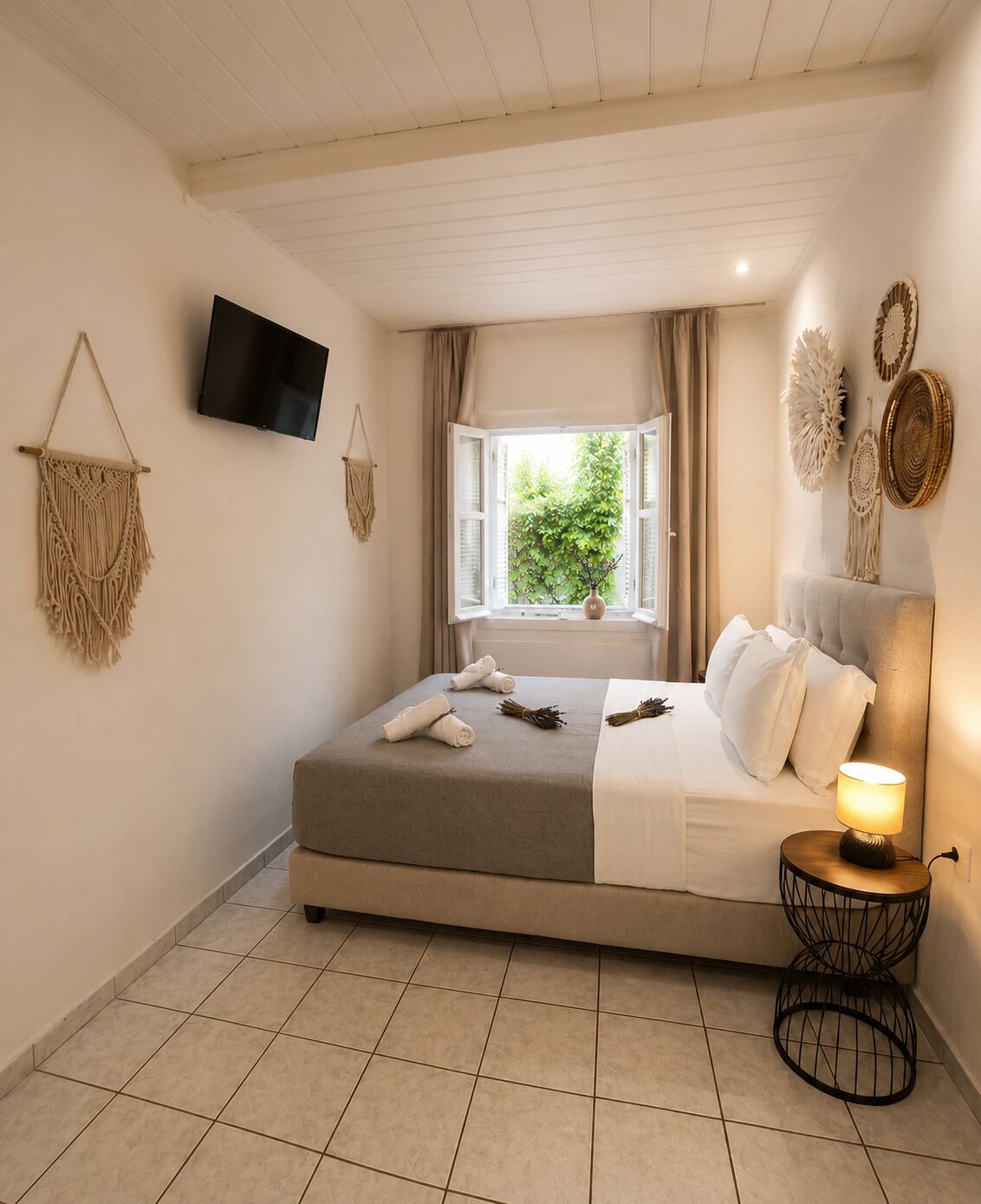 A bedroom with a bed, a bedside lamp, a TV, a window with curtains, and wall decor.